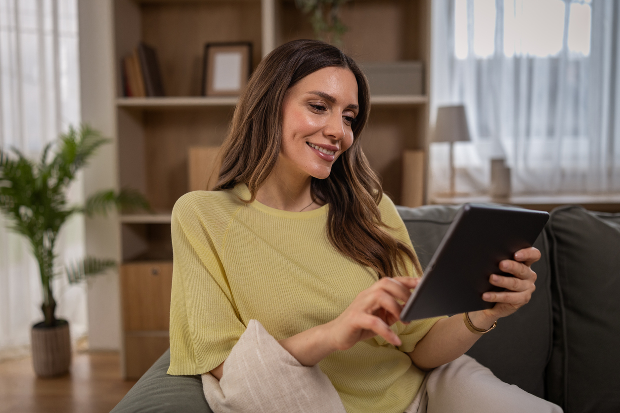 Woman sitting on couch in living room smiling while browsing digital tablet