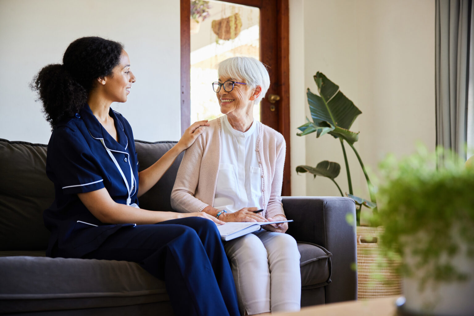 Young healthcare worker talking with a smiling senior woman while sitting together on a living room sofa during a home visit