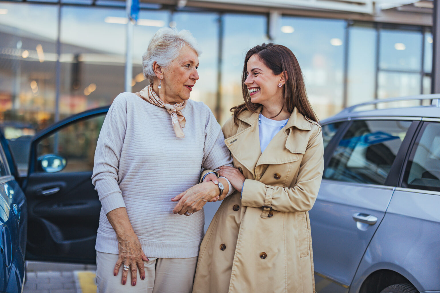 A cheerful senior woman and a young woman share a moment of connection and joy while standing outdoors near cars.